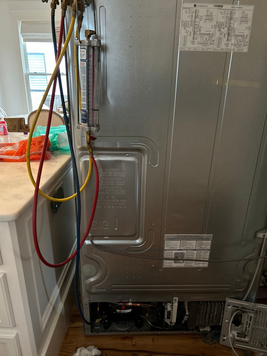 Back of an LG refrigerator with a manifold gauge set hanging from the back panel and its hoses running down to the compressor area at the bottom