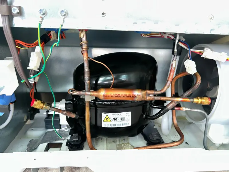 Black refrigerator compressor with copper tubing, brass fittings and wiring, seen in the open compressor compartment