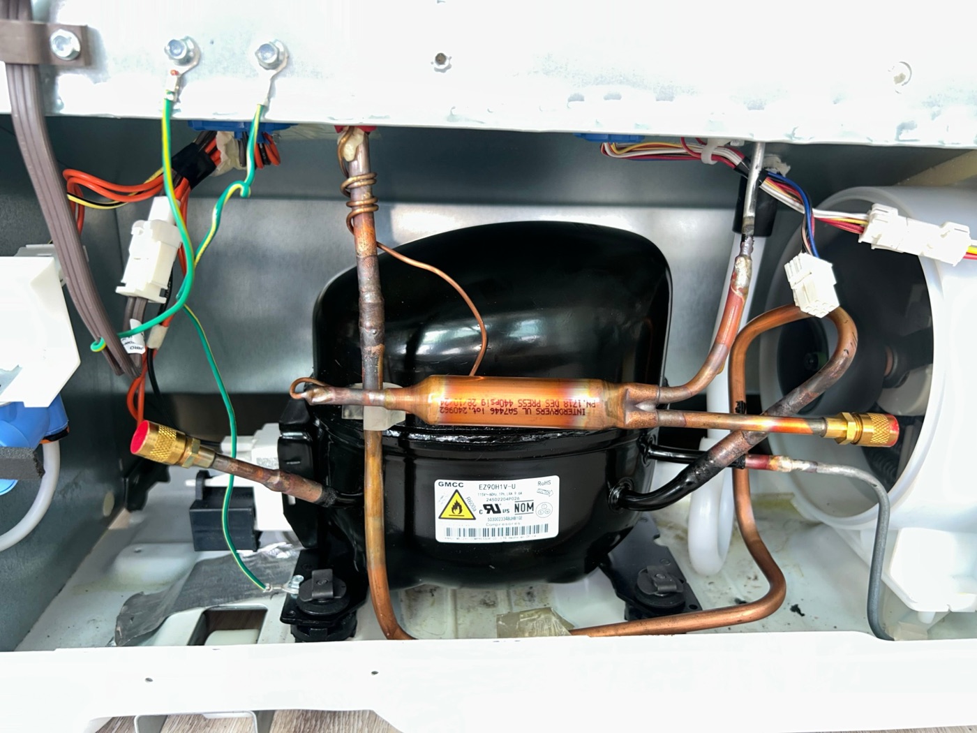 Black refrigerator compressor with copper tubing, brass fittings and wiring, seen in the open compressor compartment