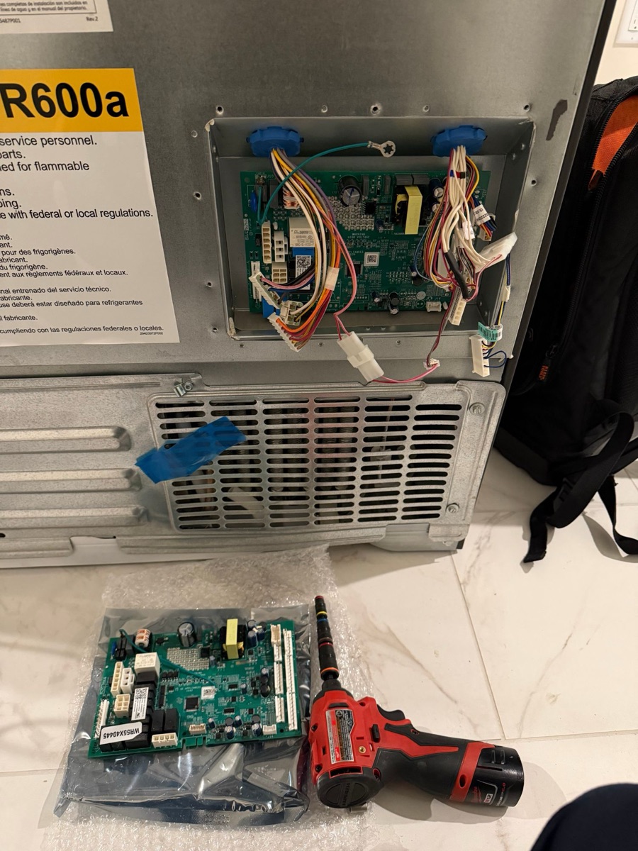 The control board in the back recess of the refrigerator with its wiring connected, and a replacement control board in its anti-static bag on the floor beside a cordless drill