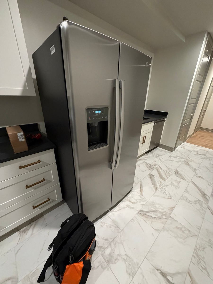 Side-by-side refrigerator in a kitchen with its control display showing zeros and a technician's tool backpack on the floor beside it